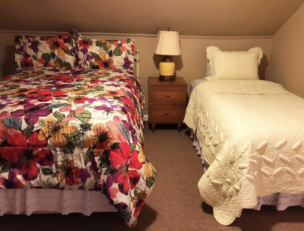 A bedroom with two beds, one with a floral bedspread and the other with a plain white bedspread. There is a nightstand with a lamp between the beds.