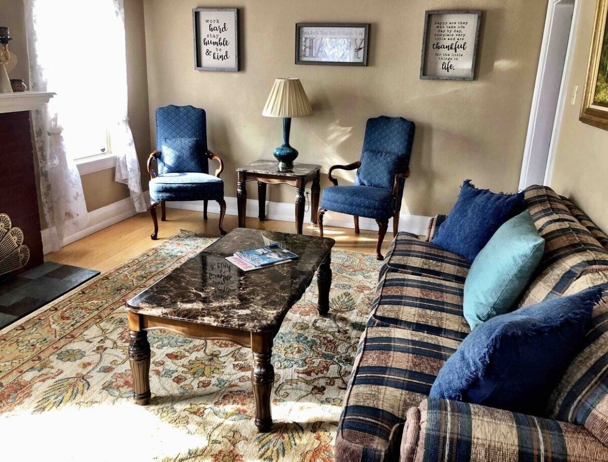 Un salon comprenant un canapé, deux fauteuils, une table basse et une lampe. Des citations encadrées ornent le mur.