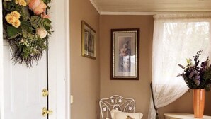 Une pièce avec une porte blanche, une chaise en métal blanc avec un coussin, une petite table sur laquelle se trouve un vase, et un tableau encadré accroché au mur.