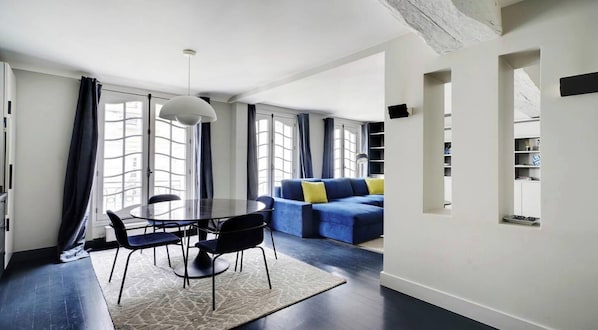 A modern living room with a dining area, featuring a round table with chairs, a blue sofa with yellow cushions, and a large window with curtains. - A PERFECT BLEND OF COMFORT & PARISIAN CHARM AT RUE SAINT SULPICE – SAINT GERMAIN (paris)