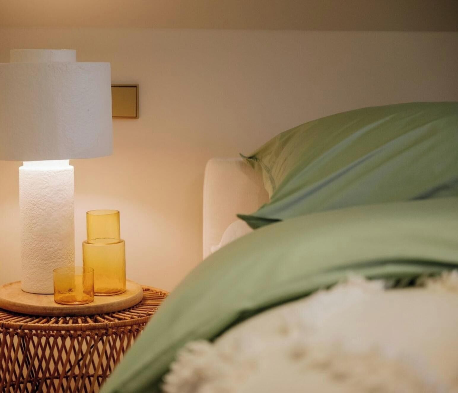 Een bed met groene lakens, een bedlamp met een witte kap en twee glazen kandelaars op een rieten nachtkastje.