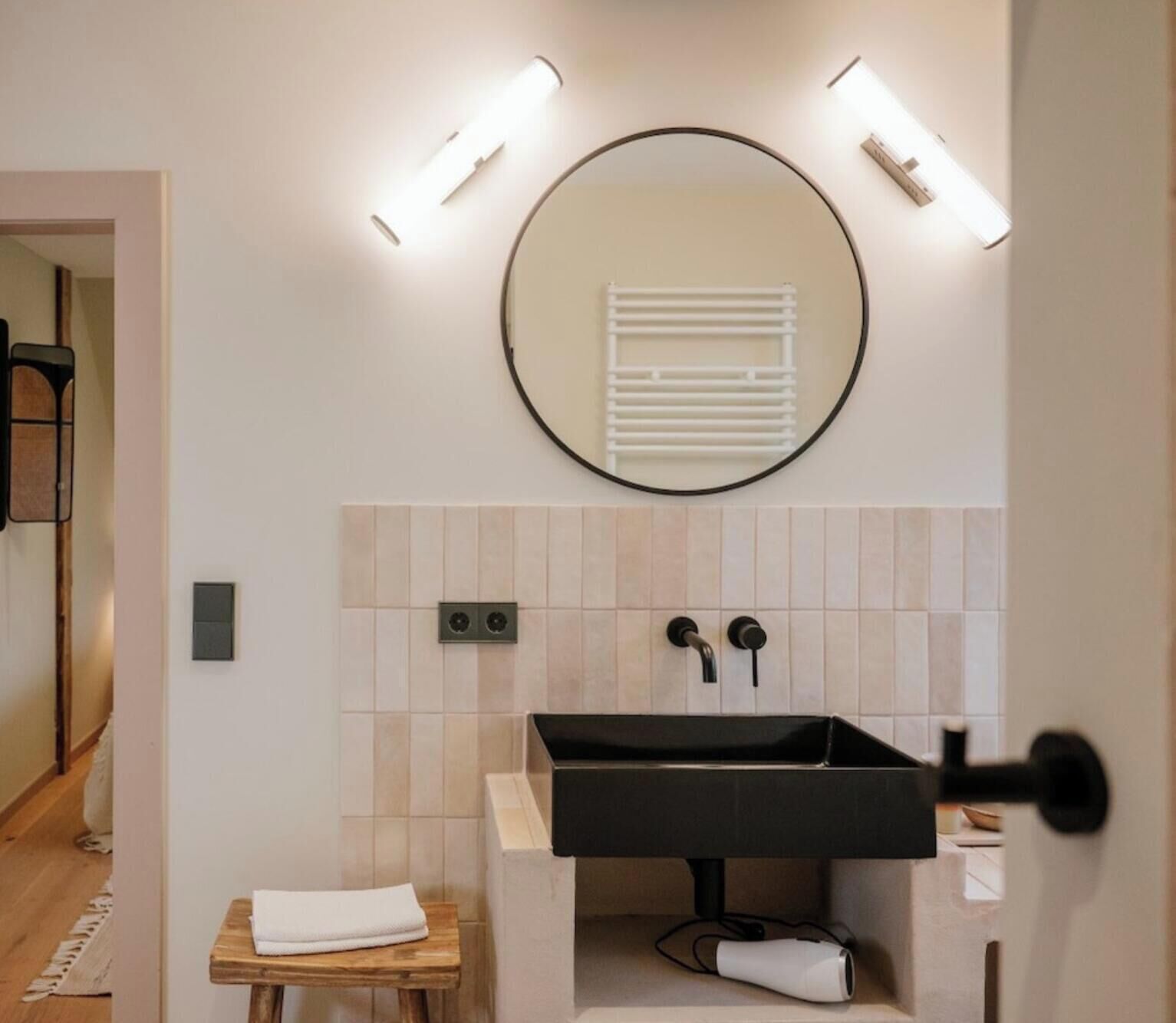 Une salle de bain avec un miroir rond, un lavabo noir et une serviette posée sur un tabouret en bois.