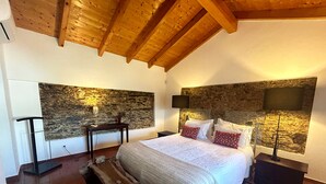 A bedroom with a stone wall, a wooden ceiling, a bed with white bedding, and a nightstand with a lamp.