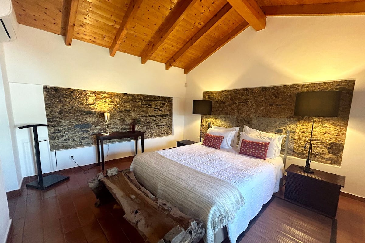 Ein Schlafzimmer mit Stehwänden, Holzbalkendecke, einem Bett mit weißer Bettwäsche und einem Nachttisch mit Lampe.