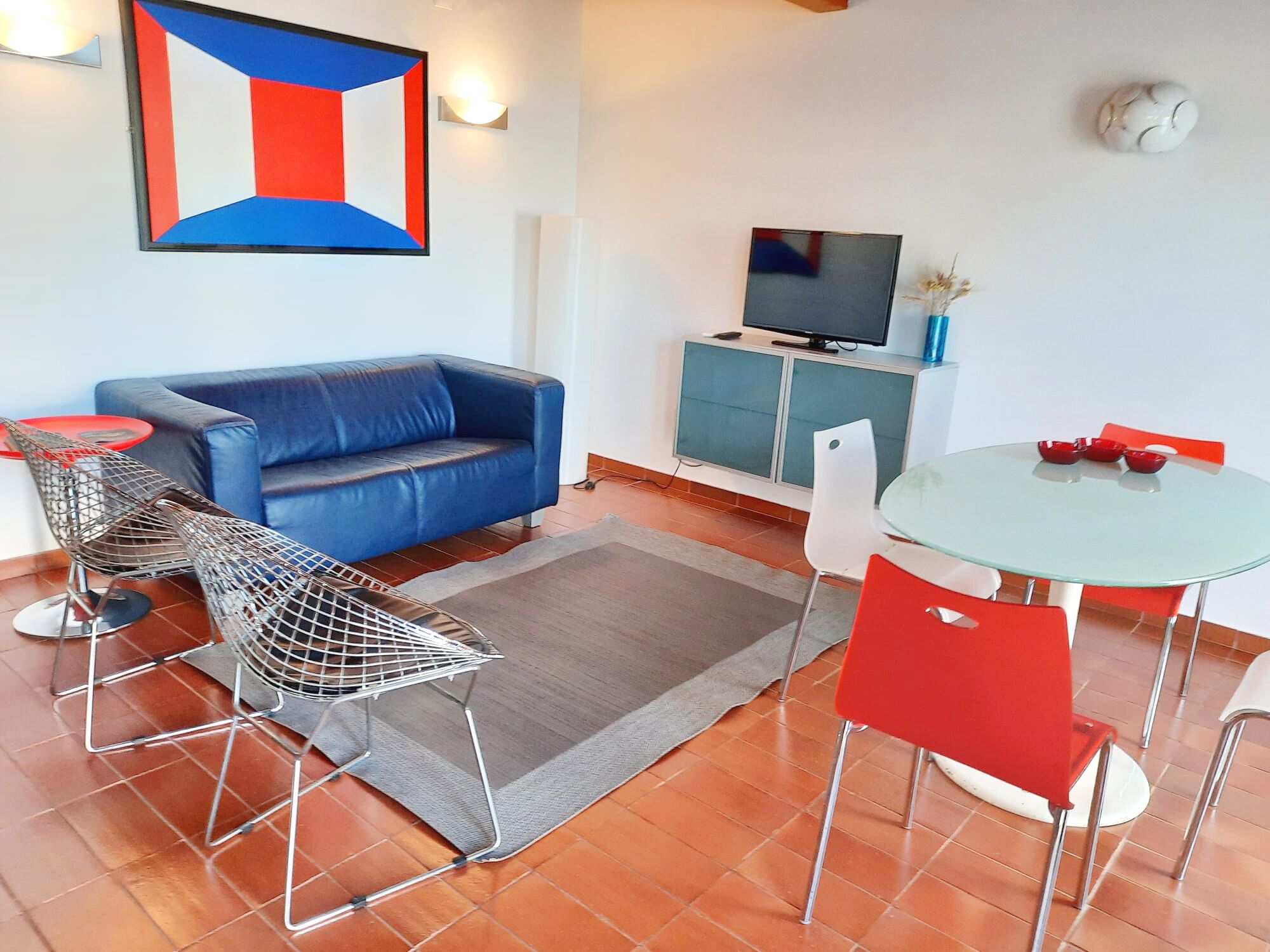 A modern living room with a blue sofa, a TV on a cabinet, a small dining table with red chairs, and a geometric artwork on the wall.