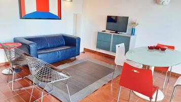 A modern living room with a blue sofa, a TV on a cabinet, a small dining table with red chairs, and a geometric artwork on the wall.