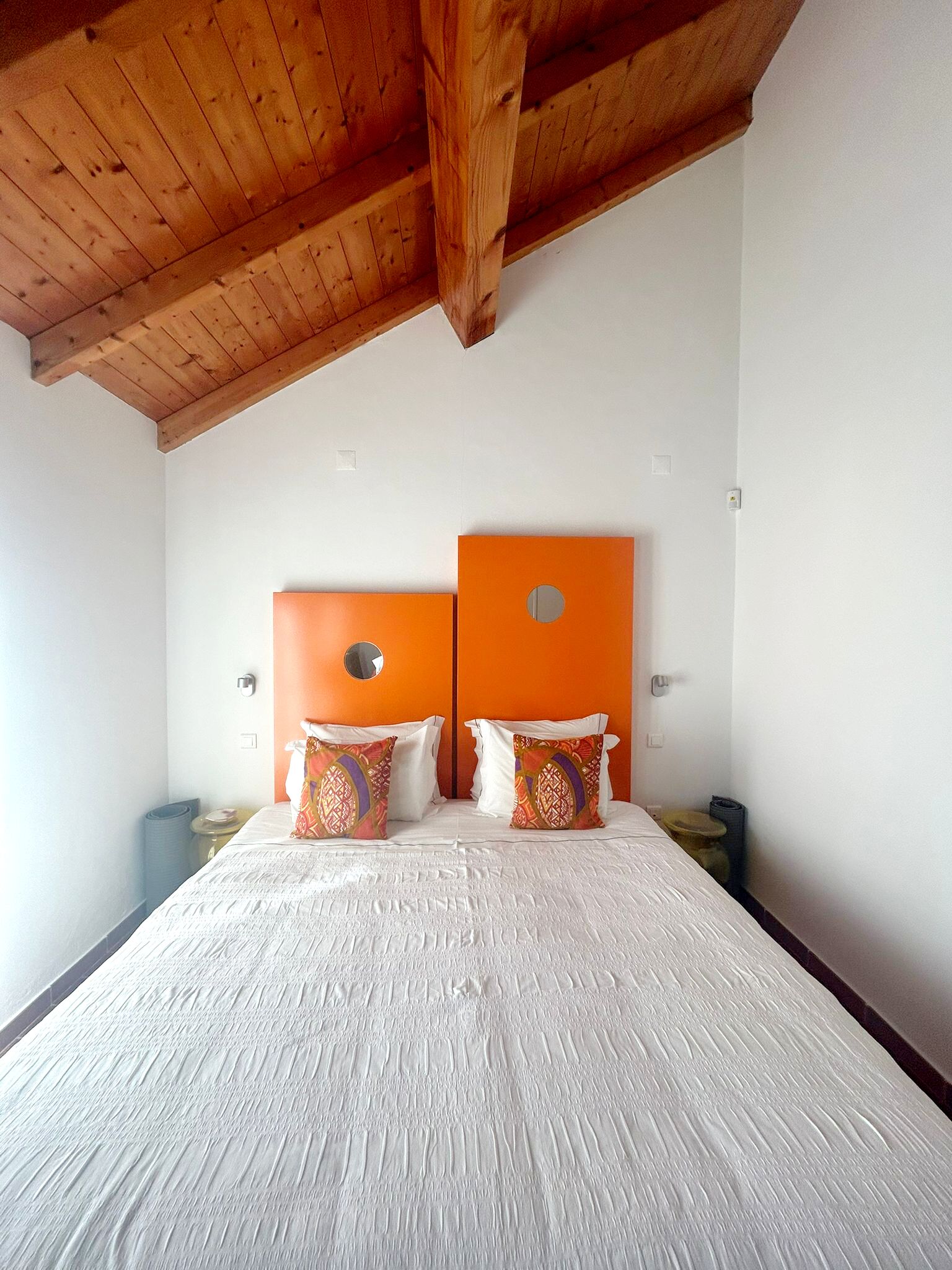 Une chambre avec un plafond en bois, deux têtes de lit orange, du linge de lit blanc et une petite table de chevet avec une lampe.
