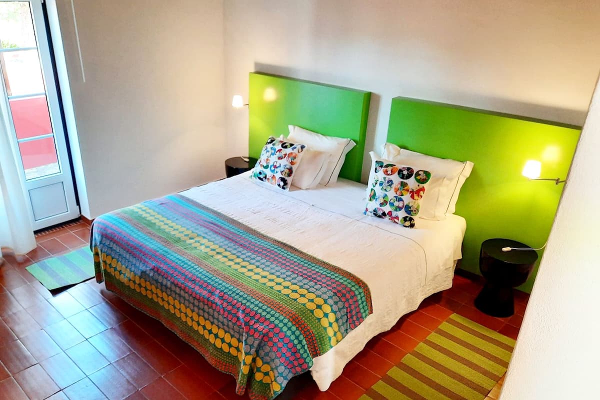 Ein Hotelzimmer mit einem großen Bett, einem grünen Kopfteil, weißer Bettwäsche, bunten Kissen und einem Streifen Teppich.