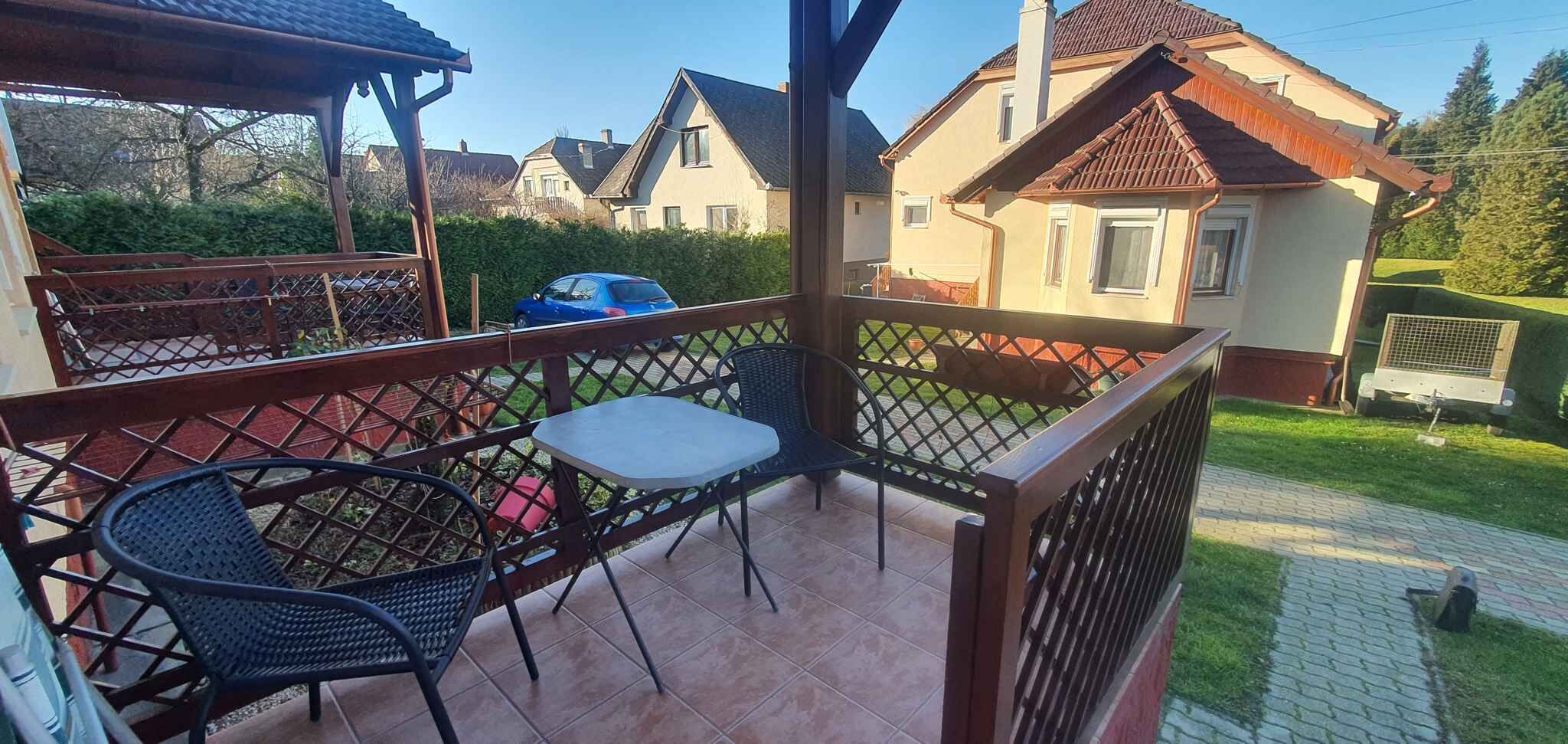 A covered outdoor patio with a table and chairs, surrounded by a wooden fence, with houses in the background.