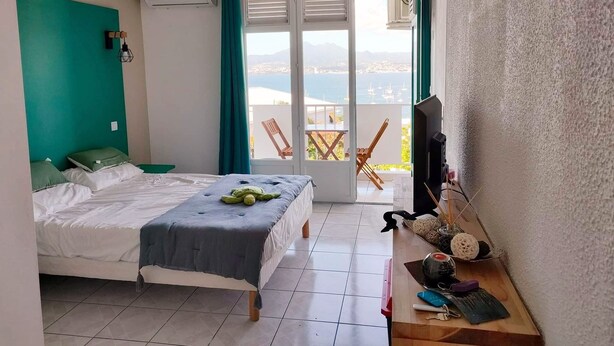 A bedroom with a bed, a desk with a TV, and a balcony with chairs. - CHAMBER ANTILLAISE (Trois ilets)