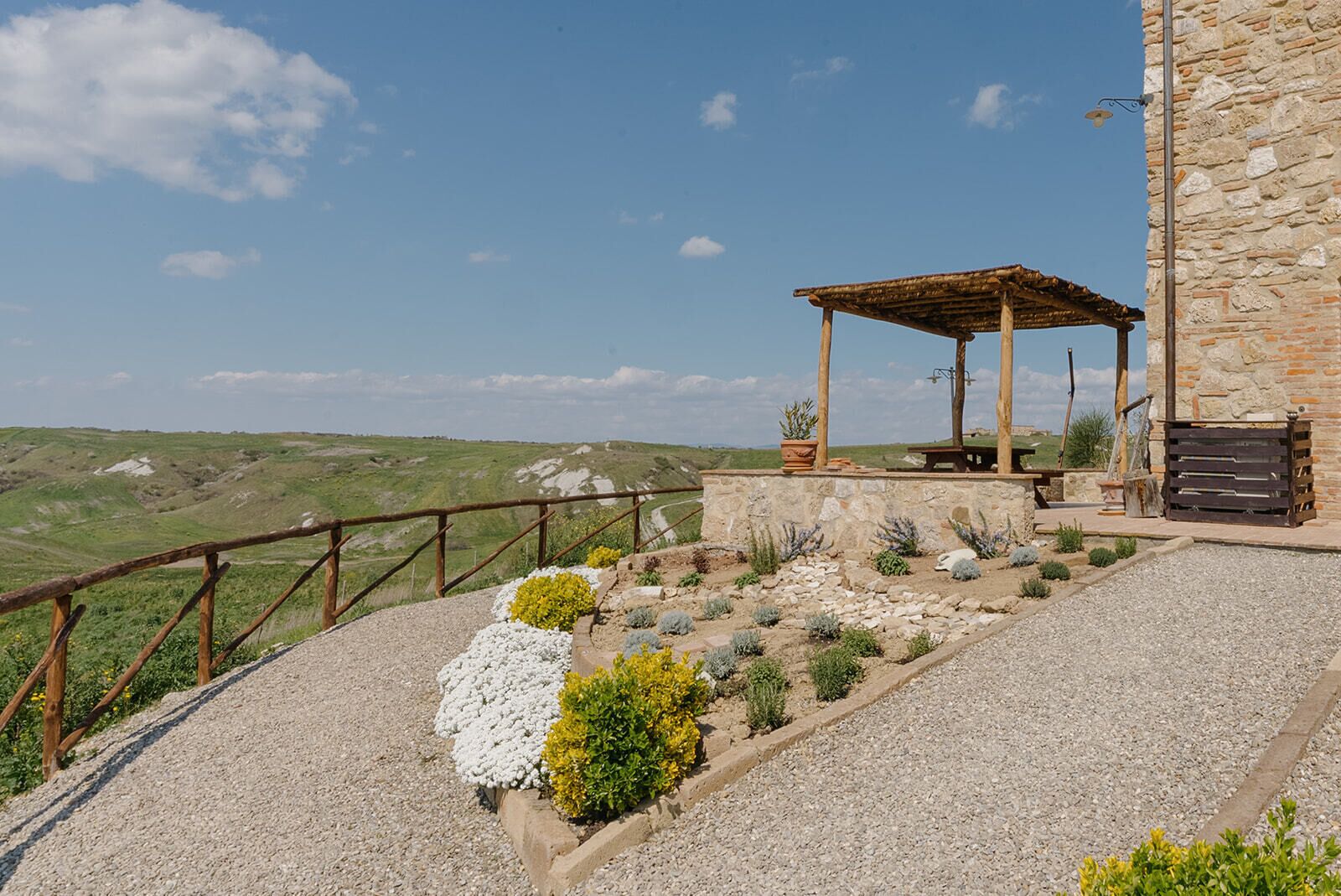 A stone building with a wooden pergola, gravel pathways, and a view of the countryside.