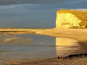 A beach with a large cliff, calm water, and a few people walking along the shore.