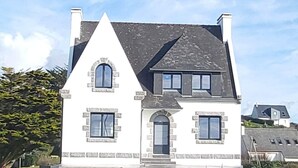 Une maison à deux étages, avec un toit à pignon, des fondations en pierre et une clôture en pierre à l’avant.
