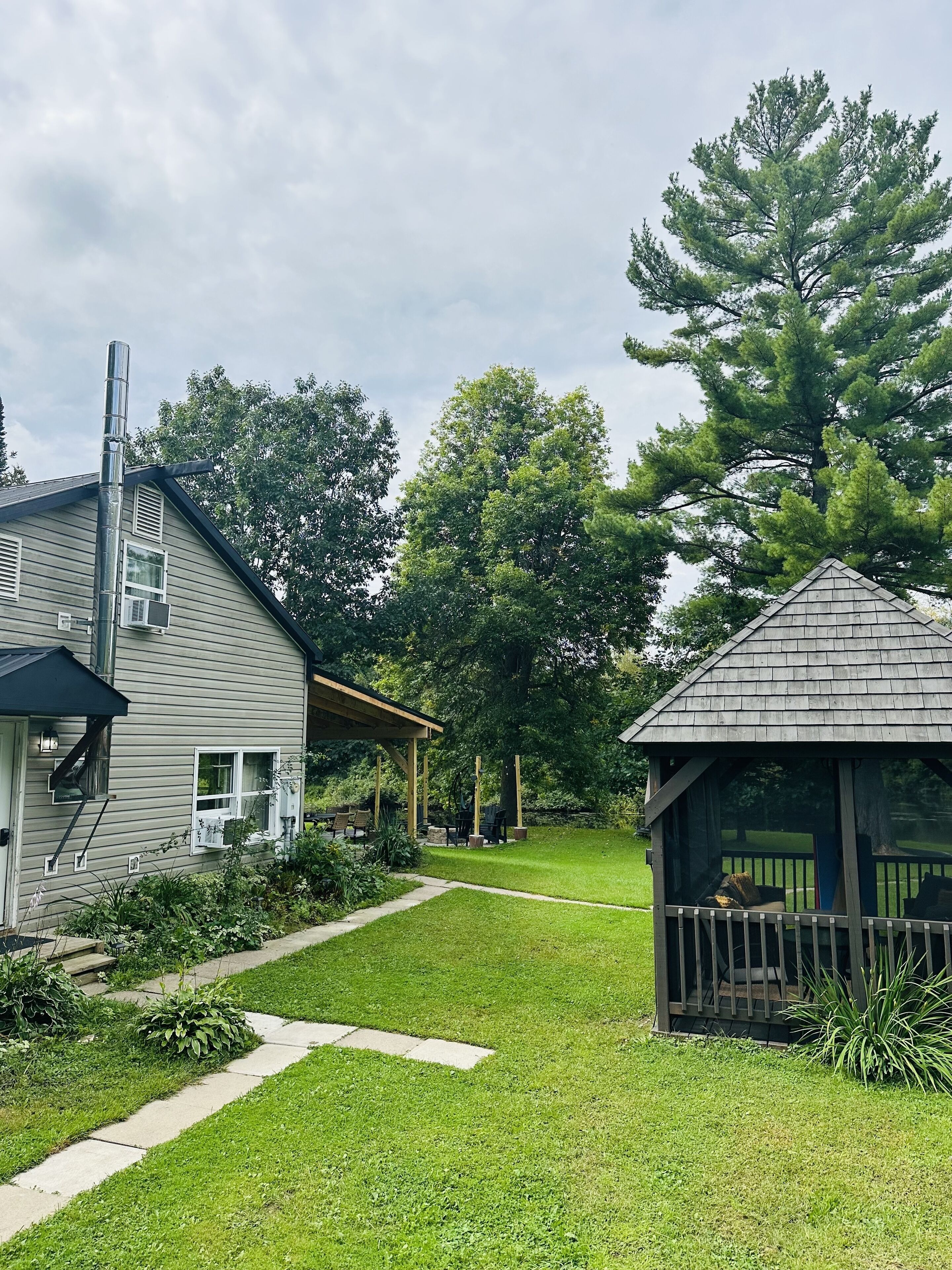 Une maison avec un patio couvert, une petite construction en bois et un grand arbre en arrière-plan.