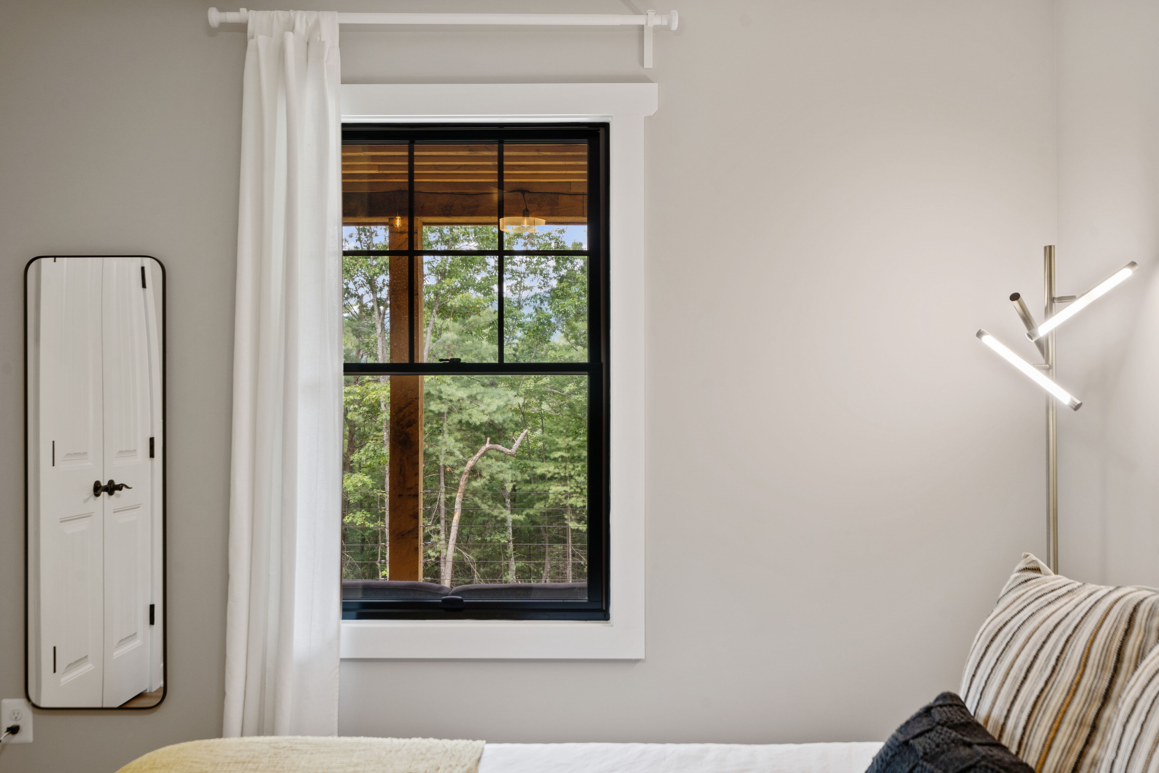 A bedroom with a large window, a bedside table, a lamp, and a wardrobe.