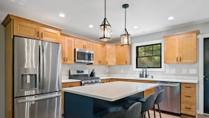 A modern kitchen with stainless steel appliances, wooden cabinets, a central island with bar stools, and pendant lighting.