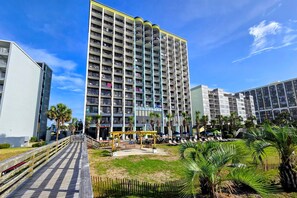 A high-rise residential building with multiple balconies, a boardwalk, and a small outdoor seating area with umbrellas.