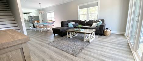 Un salon spacieux avec un canapé d’angle marron foncé, une table basse en bois et un tapis gris. On peut voir un ventilateur de plafond, et une grande fenêtre laisse entrer la lumière naturelle.