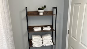 Une petite salle de bain d’angle avec une étagère en bois où sont pliées des serviettes et où est posée une petite plante.