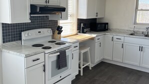 Une cuisine avec des meubles blancs, une cuisinière, un four à micro-ondes et une fenêtre avec des stores.