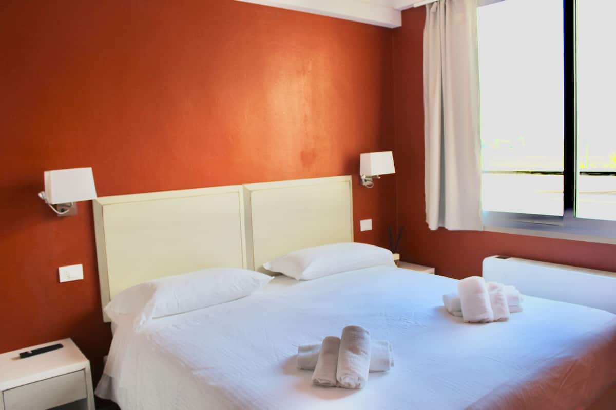 A hotel room with a large bed, white linens, two bedside lamps, and a window with curtains.