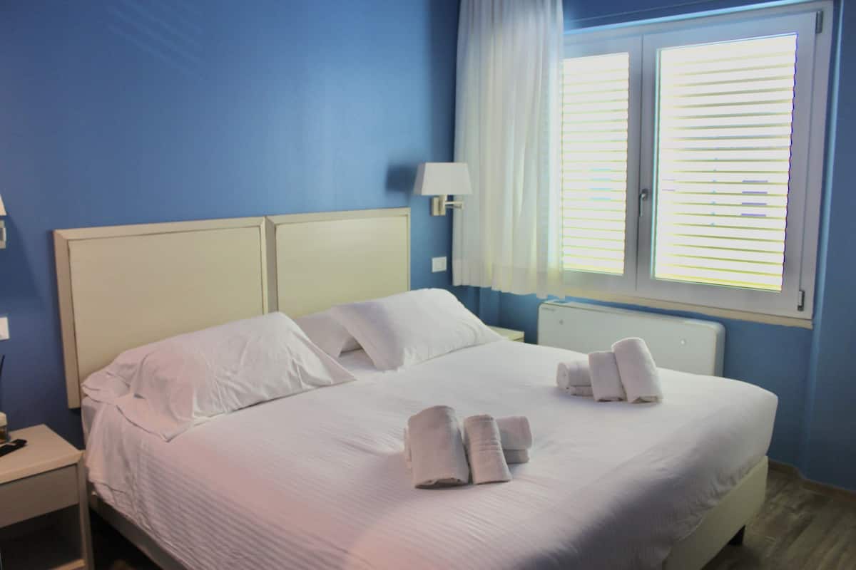 A neatly made bed with white linens and towels, a bedside table with a lamp, and a window with blinds.