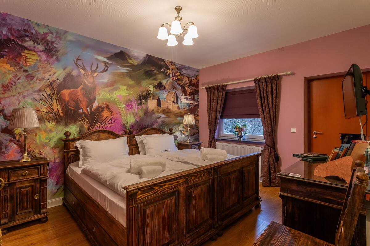 A bedroom with a large bed, a wooden dresser, a desk, a chair, a window with curtains, and a colorful mural on the wall.