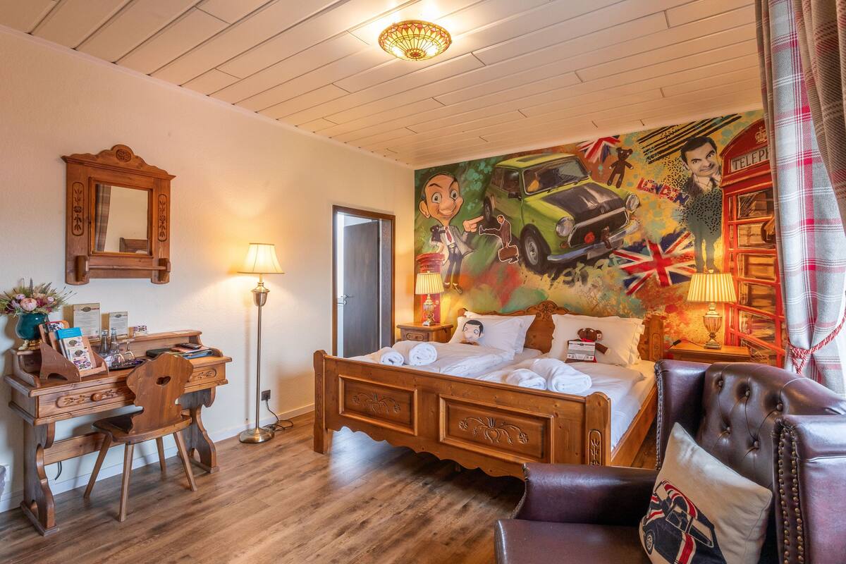 A bedroom with a wooden bed, a desk with a chair, a lamp, and a wall mural.