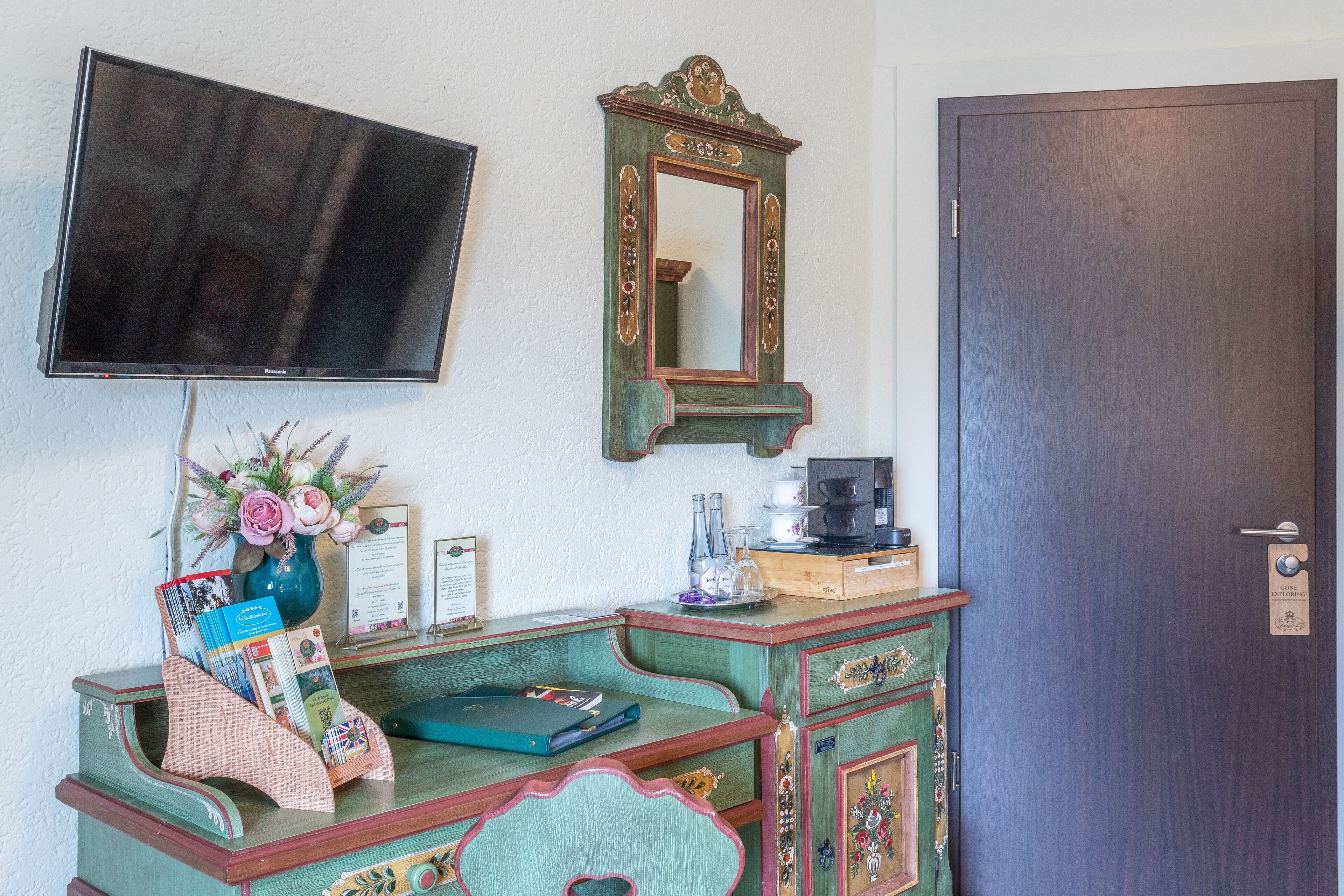 Une pièce avec un téléviseur fixé au mur, une commode en bois avec des tiroirs, un miroir et une porte avec une pancarte.