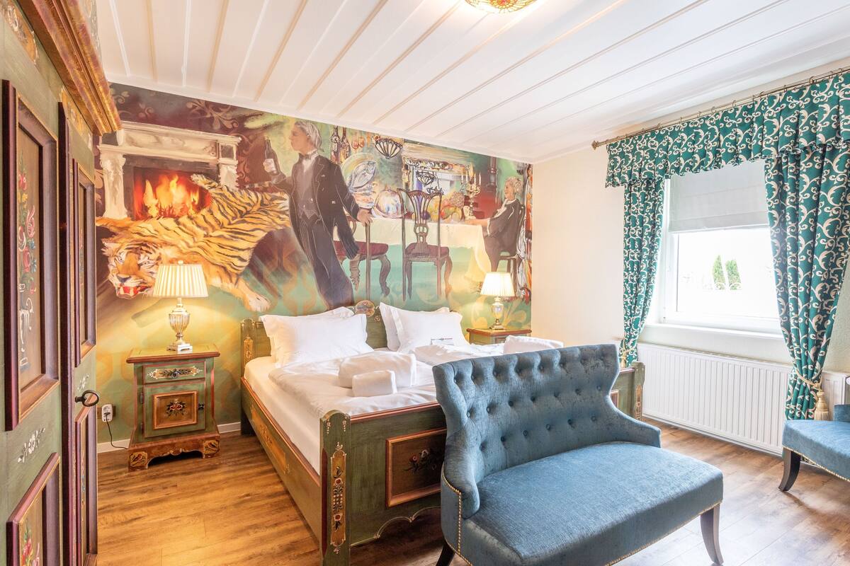 A bedroom with a large bed, a chair, a fireplace with a mural, and a window with curtains.