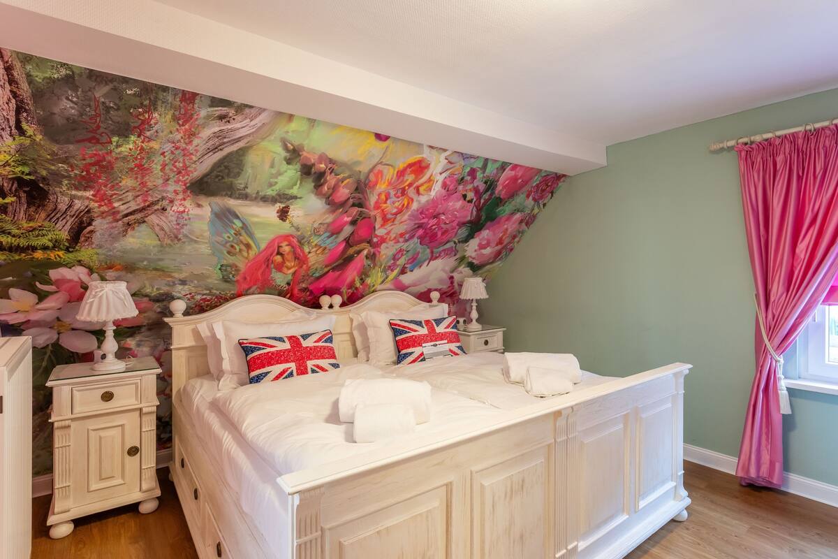 A bedroom with a large floral mural on the wall, a white bed with British flags on pillows, and a pink curtain.