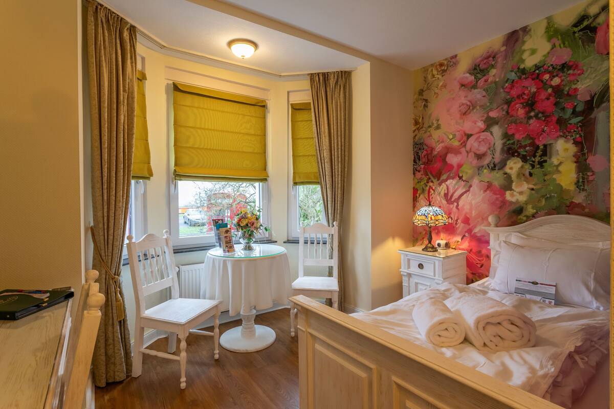 A hotel room with a bed, a round table with chairs, a window with curtains, and a floral wallpaper.