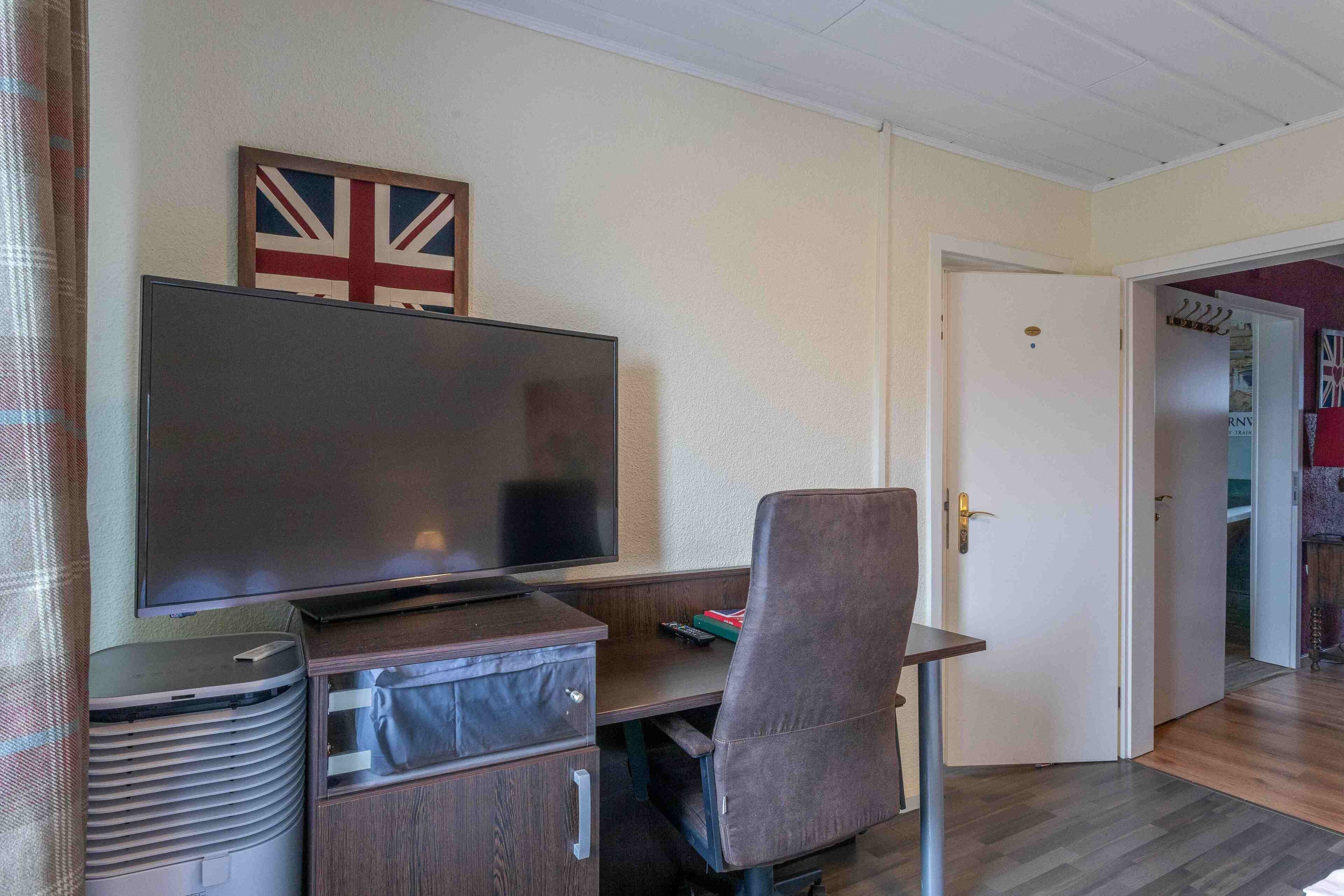 Una habitación con una televisión de pantalla plana montada en un mueble de madera, una silla de oficina gris y una puerta que da a otra habitación.