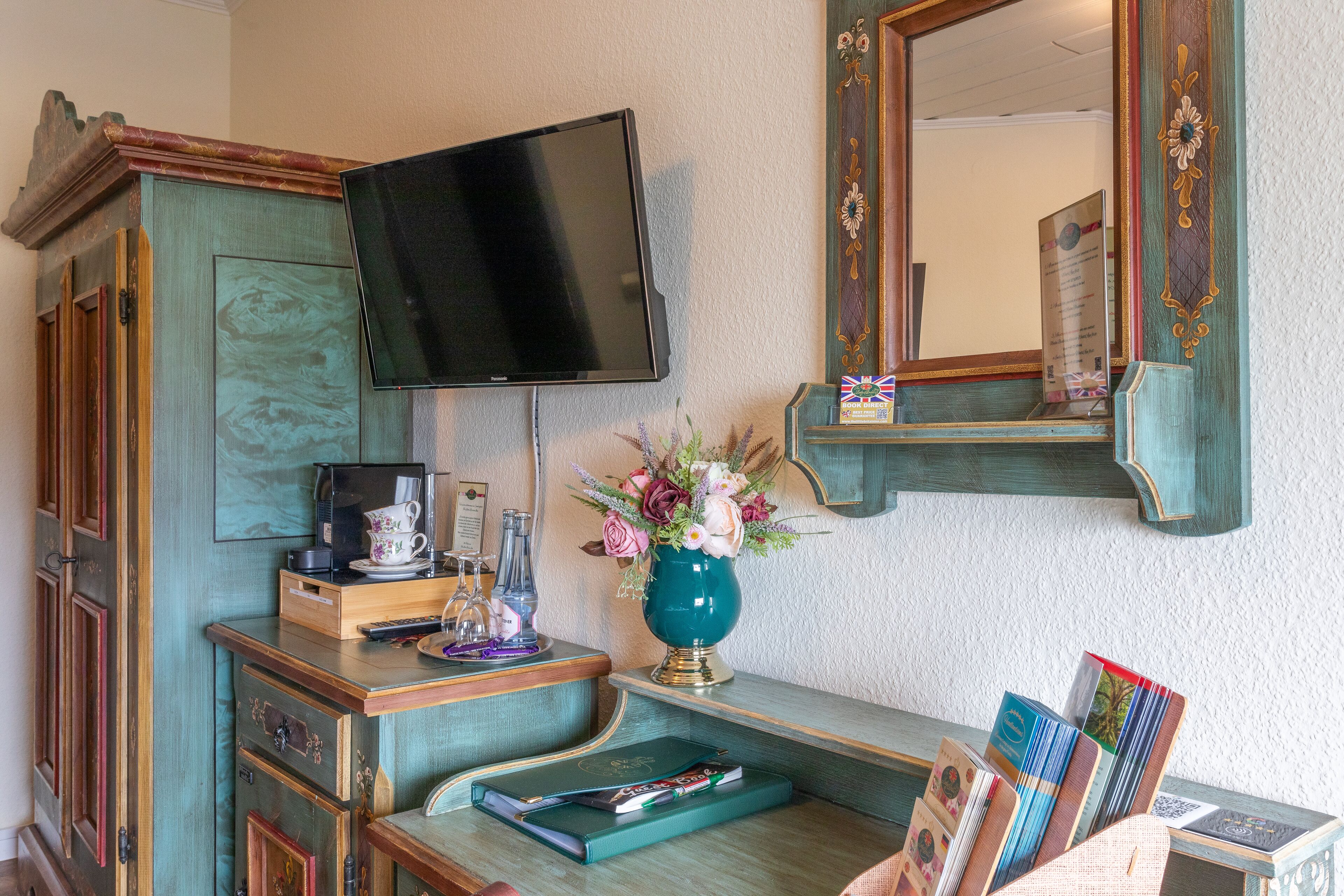 Ein Zimmer mit an der Wand montiertem Fernseher, einer Holzkabine mit Schubladen, einem kleinen Tisch mit Blumen in einer Vase und einem Spiegel über dem Tisch.