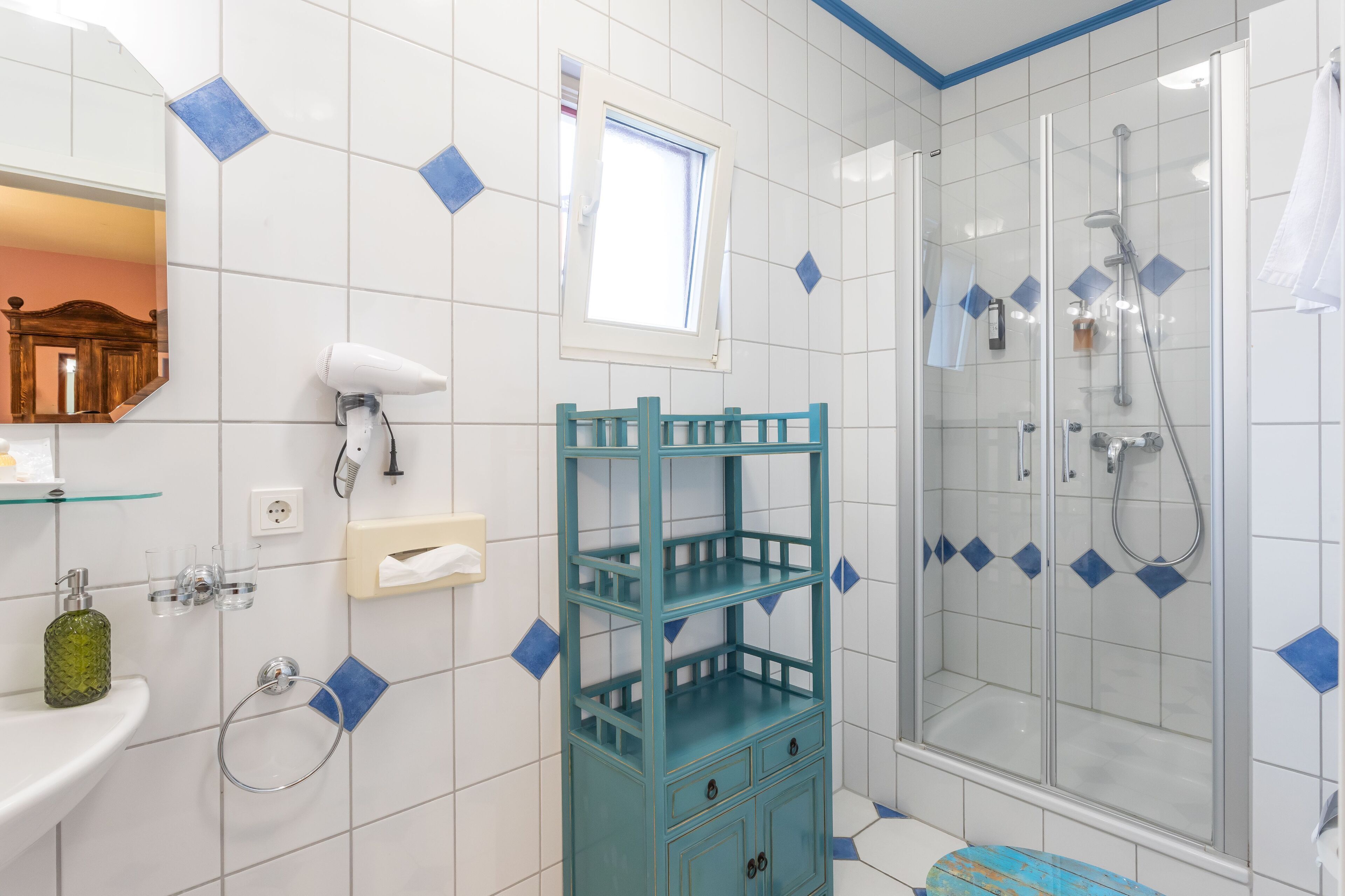 Une salle de bain avec une douche à parois de verre, un lavabo blanc avec un miroir et un meuble de rangement de couleur turquoise.