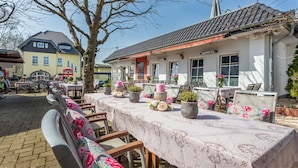 An outdoor dining area with tables set for guests, surrounded by a building with a tiled roof and a flower-adorned fence.