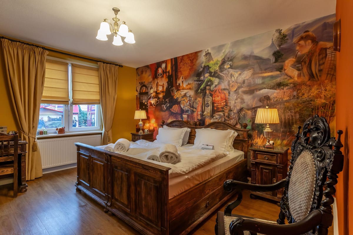 A bedroom with a large bed, wooden headboard, two bedside tables with lamps, a window with curtains, and a mural on the wall.