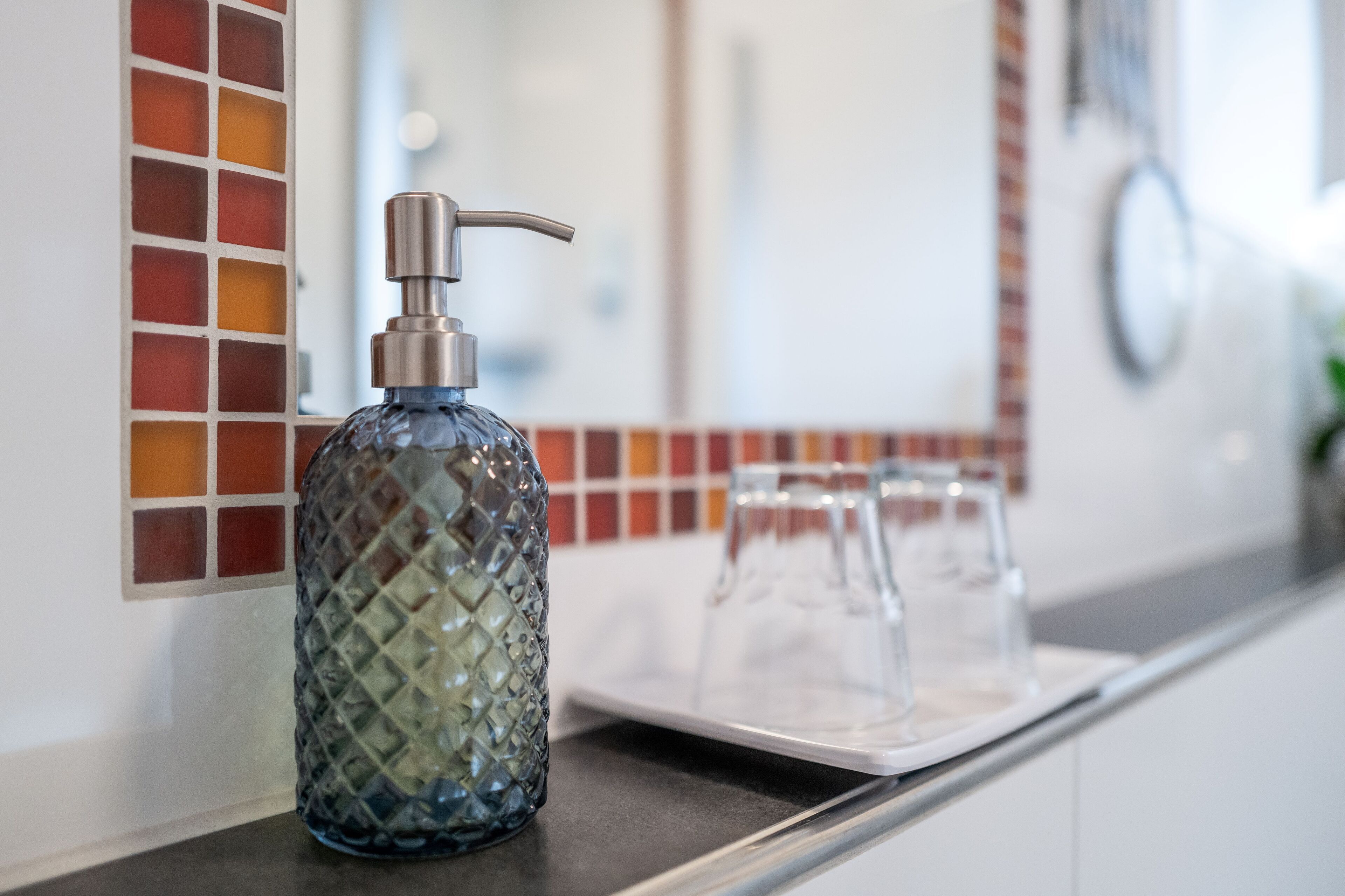 A bathroom with a glass soap dispenser on a counter, a mirror, and a tiled backsplash.