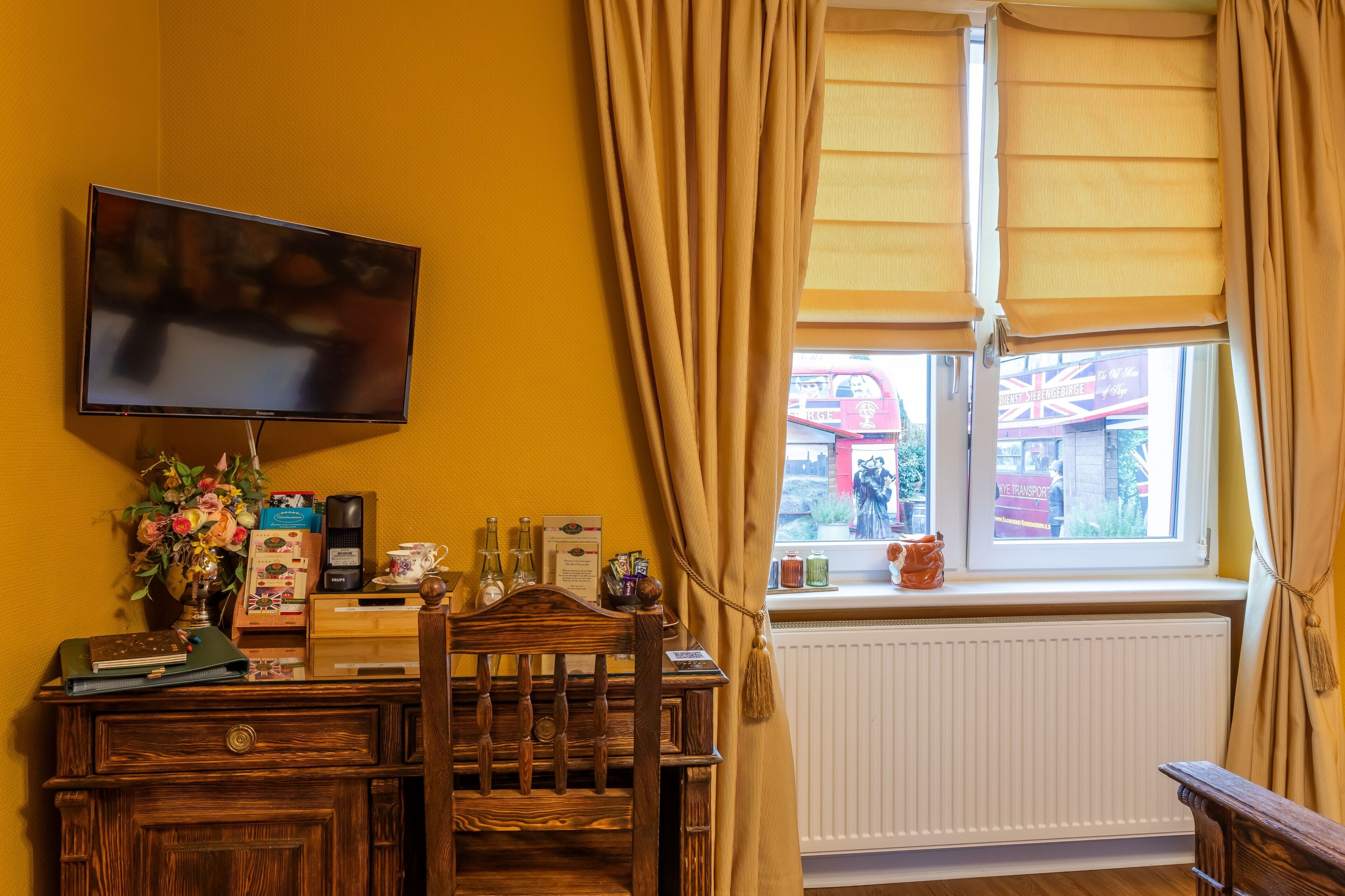 A room with a TV mounted on the wall, a wooden desk with various items, a chair, and a window with curtains.
