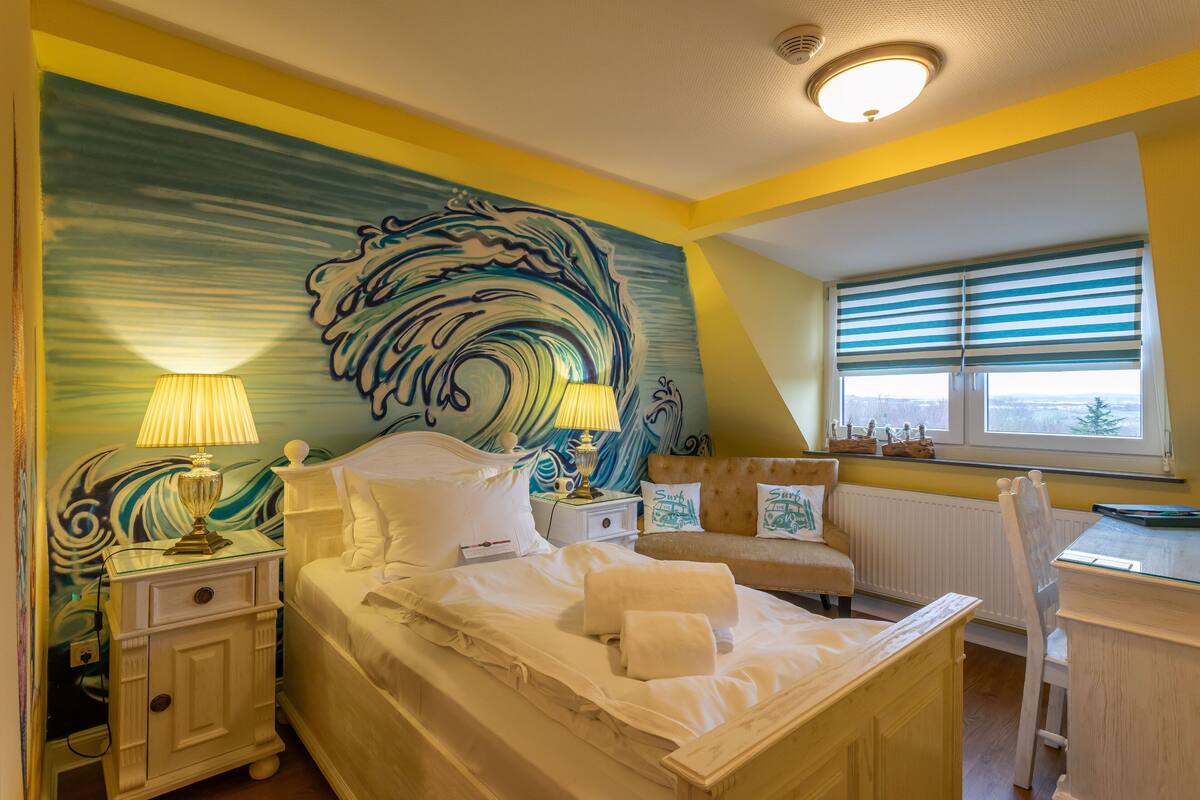 A bedroom with a bed, bedside tables, a nightstand, a window with blinds, and a view of the outside.