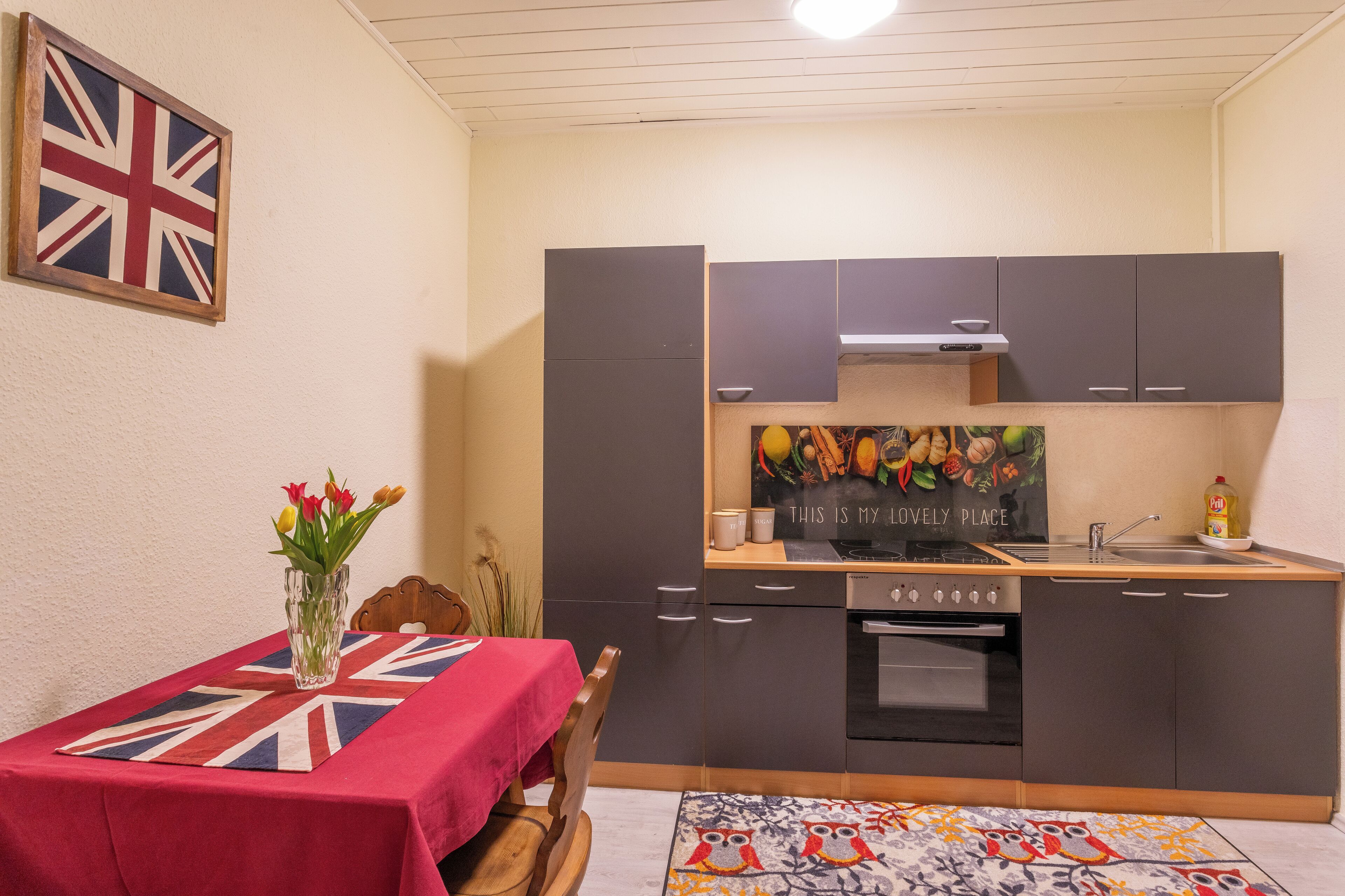 A compact kitchen with dark cabinets, a central oven, and a small countertop. A table with a Union Jack tablecloth and a vase of flowers is set against the wall.