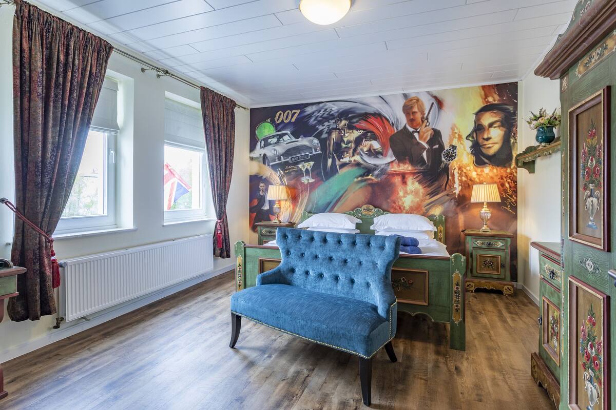 A bedroom with a large bed, a blue upholstered chair, a mural on the wall, and a window with curtains.