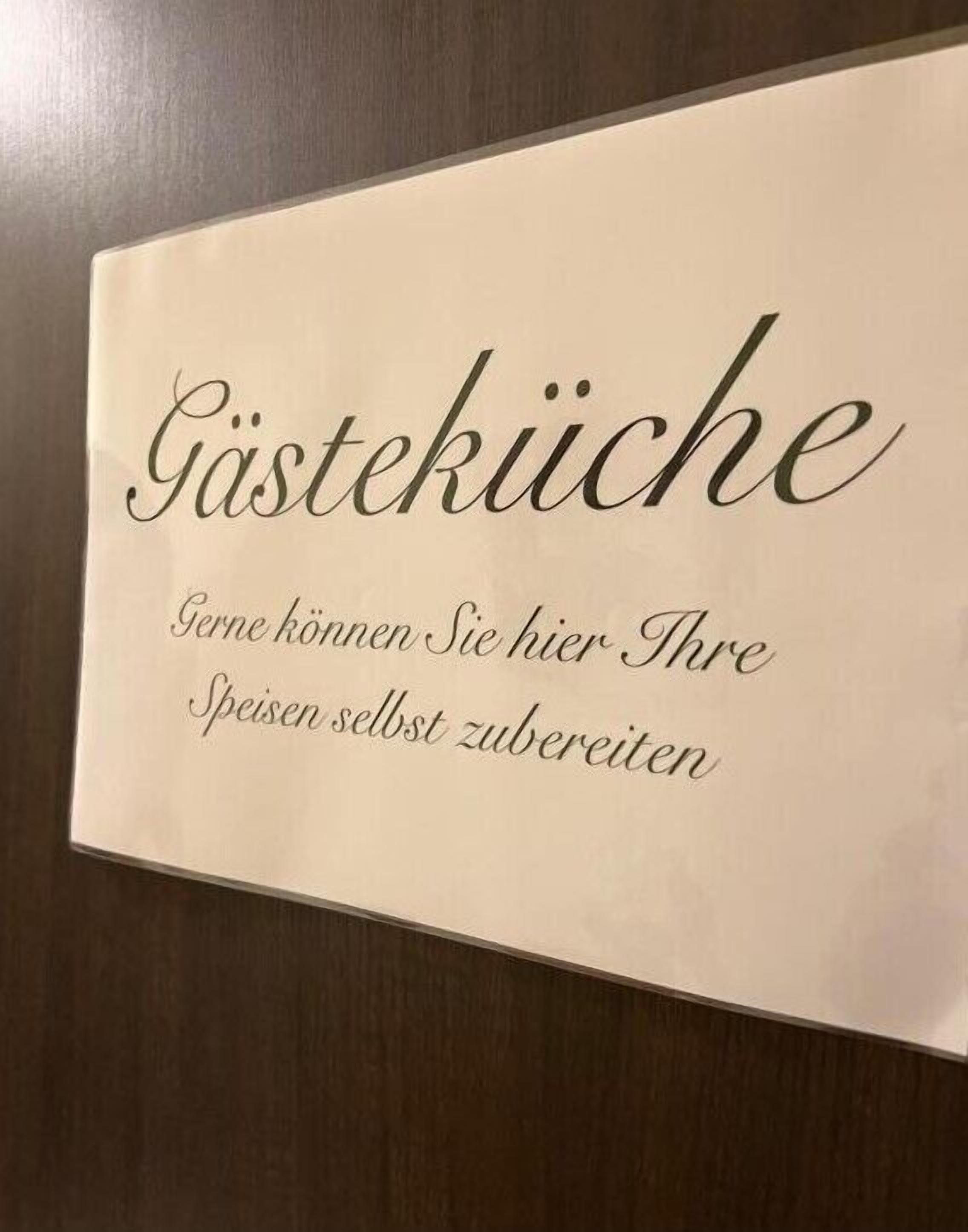 Ein Schild mit dem Text "Gästeküche" und "Gerne können Sie hier Ihre Speisen selbst vorbereiten" vor beige Hintergrund.