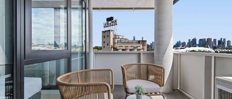 Een balkon met twee rieten stoelen, een klein rond tafeltje met een plant, en uitzicht op een stadsgezicht met een bouwplaats op de achtergrond.