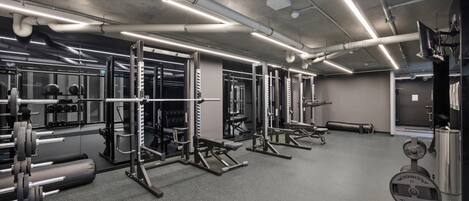 A gym with weightlifting equipment, mirrors, and a grey carpeted floor.