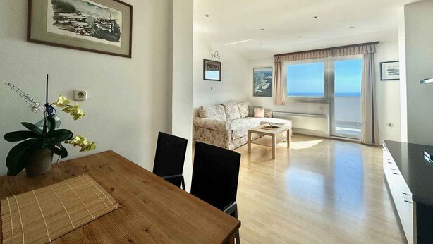 Een ruime woonkamer met een houten eettafel, een bank en uitzicht op zee door grote ramen.