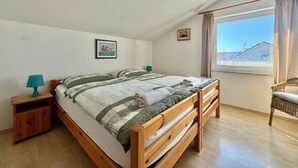 Een slaapkamer met een houten bed, een nachtkastje, een stoel, een raam met uitzicht en een ingelijste foto aan de muur.