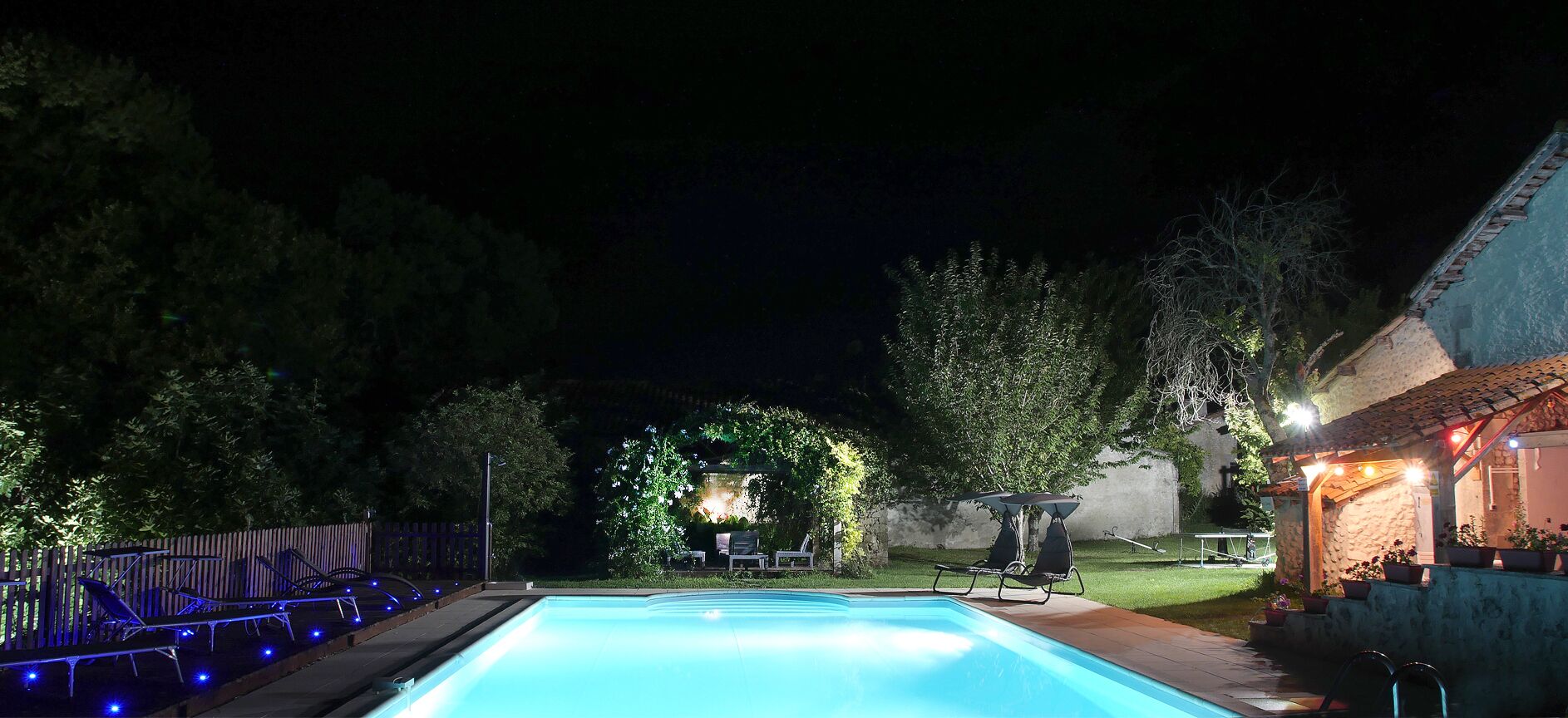 A swimming pool at night with surrounding lounge chairs and a small building with lights.