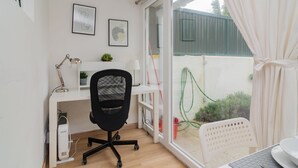 Ein moderner Arbeitsplatz mit Schreibtisch, Stuhl und Computer. Zwei gerahmte Bilder hängen an der Wand, daneben ein Blumentopf und ein Blick ins Freie durch eine Glastür.