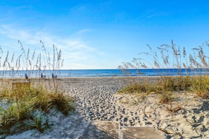 Une plage de sable fin, bordée d’herbes hautes et surplombée d’un ciel bleu clair.